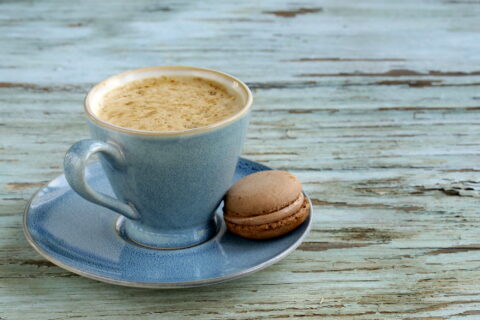 Das Bild zeigt eine Kaffeetasse, die auf einer Untertasse steht und mit Kaffee gefüllt ist. Auf der Untertasse liegt ein Macaron.