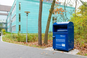 Das Bild zeigt den blauen Abfallcontainer für Altmedikamente, der vor dem Gebäude Chemikum der FAU steht.