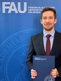 Simon Hamman vor einem Poster mit FAU-Logo, in der Hand hält er seine Ernennungsurkunde zum Professor
