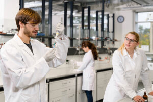 Das Foto zeigt einen Mann und zwei Frauen im Labor im Chemikum. Der Mann steht im Vordergrund und hält eine Pipette in der Hand. Die Frau rechts notiert sich etwas auf einem Schreibblock. Die Frau im Hintergrund pipettiert ebenfalls.