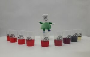 Das Foto zeigt mehrere Becher, in denen eine rote Flüssigkeit ist. Darüber schwebt eine kleine menschenähnliche Figur mit grünem Kittel.