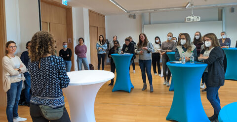 Das Foto zeigt einen Seminarraum im Chemikum. Es sind verschiedene Stehtische aufgestellt mit weißen und blauen Überzügen, an denen Studentinnen stehen und interessiert zuhören. Im Vordergrund steht Prof. Young mit dem Rücken zur Fotografin und berichtet über ihren akademischen Werdegang. Alle sind leger gekleidet in Jeans, Pullover u. ä., ein paar der Studentinnen tragen einen Mund-Nase-Schutz und haben Getränkeflaschen vor sich stehen.