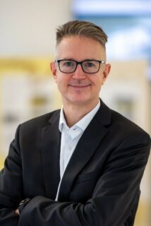 Portraitfoto von Prof. Dr. Jörg Libuda. Herr Libuda hat kurzes dunkles Haar, das an den Seiten rasiert ist. er trägt eine dunkle Brille, ein schwarzes Jackett und ein weißes Hemd. Er lächelt in die Kamera und hat die Arme dabei verschränkt vor der Brust. Der helle Hintergrund des Fotos ist verschwommen.