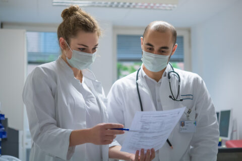 Das Foto zeigt links eine junge Pharmazie-Studentin, gekleidet in weißem Kittel und Mundschutz, und rechts einen Arzt gekleidet in weißem Kittel, Mundschutz und mit Stethoskop um den Hals gelegt. Die Studentin hält ein Blatt Papier mit Medikamentenplan in der Hand, das sie dem Arzt zeigt. Der Arzt trägt ein Namensschild, auf dem das Logo des Universitätsklinikums Erlangen zu erkennen ist.