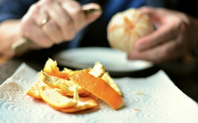 Das Foto zeigt zwei Hände, die mit einem Messer eine Orange schälen. Im Vordergrund liegen die Orangenschalen auf einem Papiertuch.